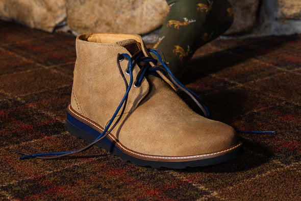 Tan suede chukka boot with navy laces on a plaid rug