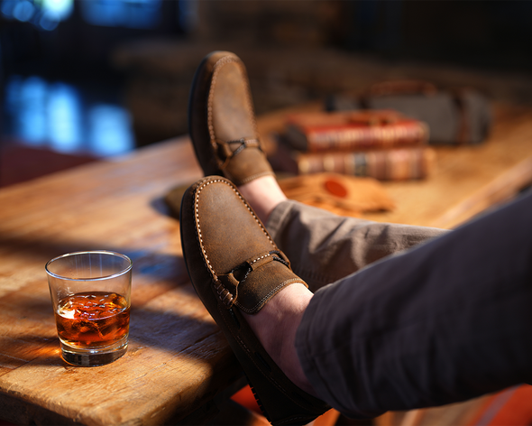 Men's shoes rested on wood table with a drink in a glass cup.